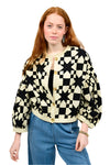 Person wearing a black and white patterned cardigan with blue jeans on a plain background