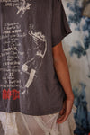 Person wearing a dark t-shirt with graphic design and text, standing against a blurred background.
