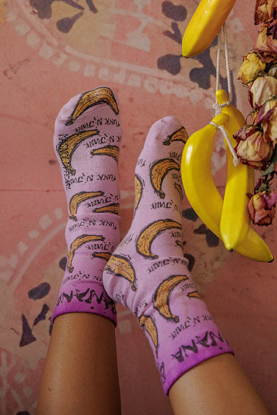 Purple socks with banana pattern worn by a person, with a pink background and yellow scissors.