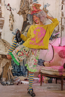 Person in colorful outfit with yellow sweatshirt and floral skirt in eclectic room.