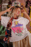 Person wearing a colorful hat and graphic t-shirt with a floral scarf, standing against a decorative background.