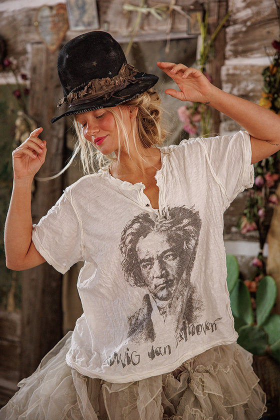 Person wearing a white t-shirt with a printed face design and a black hat, standing in front of a rustic stone wall.