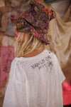 Person wearing a patterned hat and white shirt with text, standing in a blurred outdoor setting.
