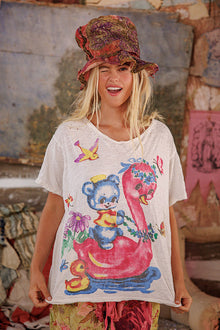  Woman wearing a white t-shirt with colorful graphics and a patterned hat in a rustic setting.