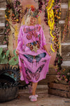 Woman in a pink dress with butterfly designs standing in a decorative setting with flowers and plants.