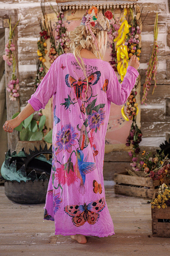 Woman wearing a pink floral and butterfly patterned dress in a rustic setting with hanging flowers.