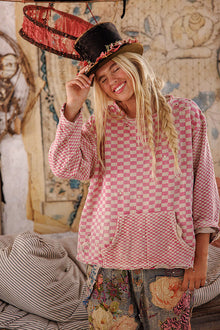  Woman wearing a pink checkered shirt and floral pants, holding a top hat in a room with decorative walls.