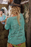 Person wearing a teal t-shirt with text, standing in a rustic setting.