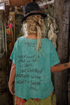 Person wearing a teal t-shirt with text, standing in a rustic setting.