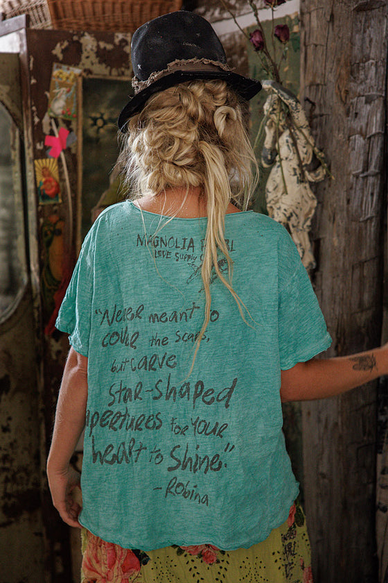 Person wearing a teal t-shirt with text, standing in a rustic setting.