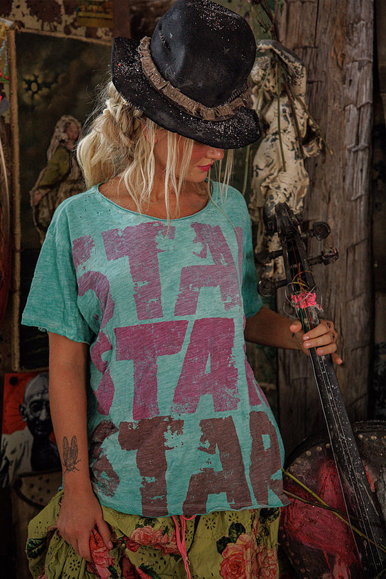 Person wearing a teal t-shirt with pink and red text, holding a bow and arrow in a rustic setting.