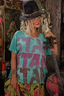  Person wearing a teal t-shirt with 'STAR' pattern, holding a guitar in a rustic setting.