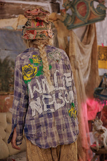  Person wearing a colorful plaid shirt with 'Willie Nelson' text, standing in a room with artistic decor.