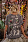 Person wearing a dark gray t-shirt with graphic designs and text, standing in a decorative setting with flowers and plants.