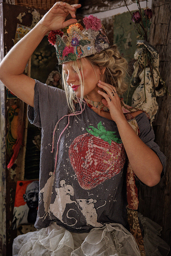 Person wearing a colorful beaded headpiece and a t-shirt with a strawberry design in a rustic setting.
